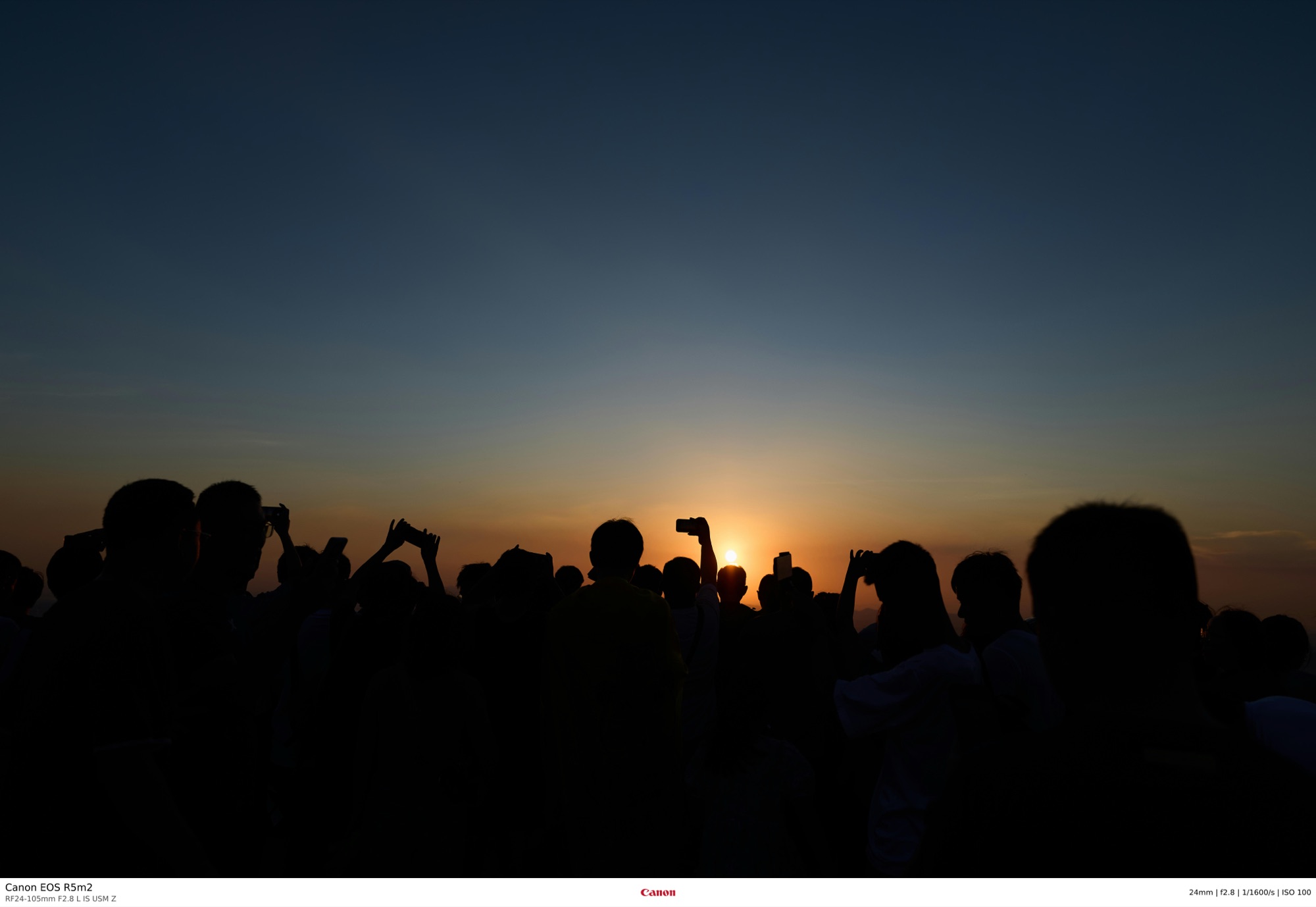 Silhouetted crowd raising phones and cameras toward the sun on a hazy mountaintop at sunrise